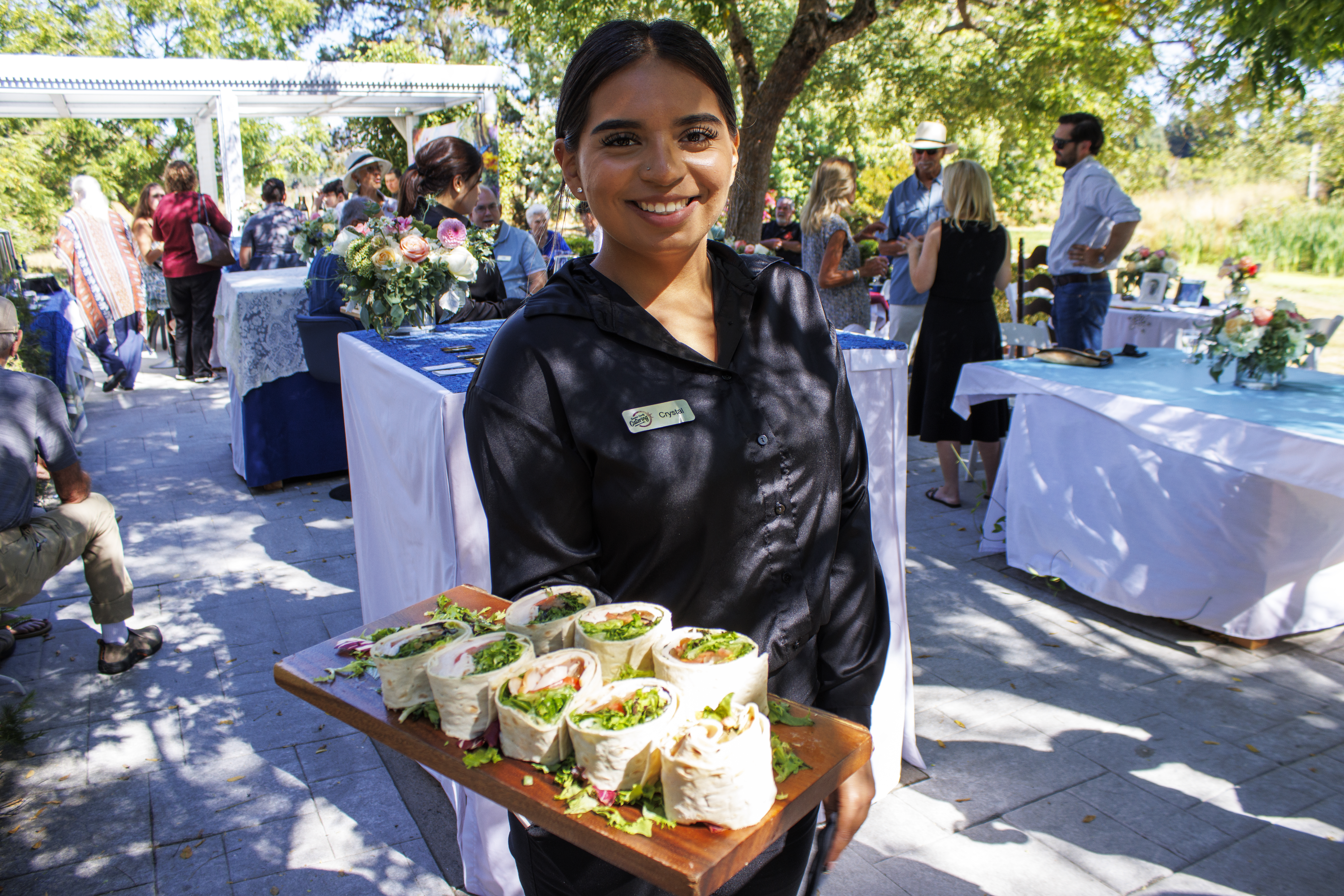 A young woman in a black silk chef's uniform carries a tray of pinwheel wraps decorated with lettuce and tomato. The woman...