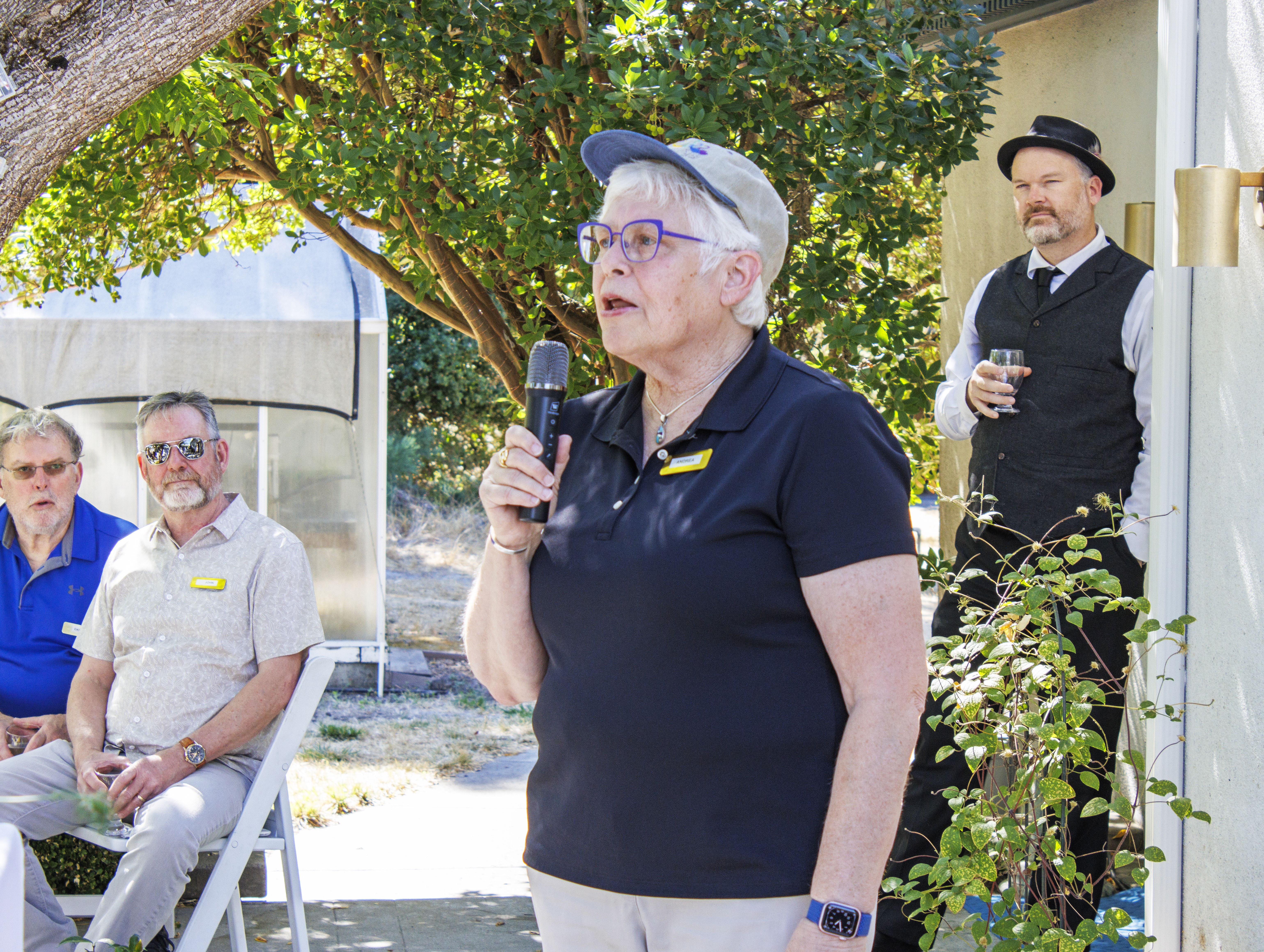 A group of older adults are gathered together, one holding a microphone. They are standing in front of a white wall with v...