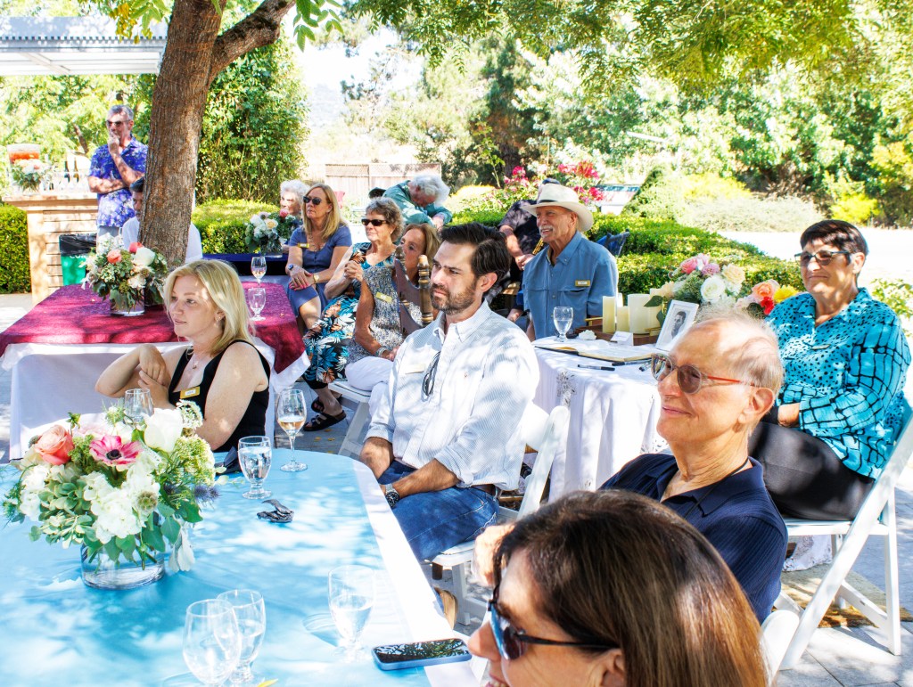 A sunny outdoor gathering of people in an outdoor setting. In the foreground, there is a table made of blue fabric covered...