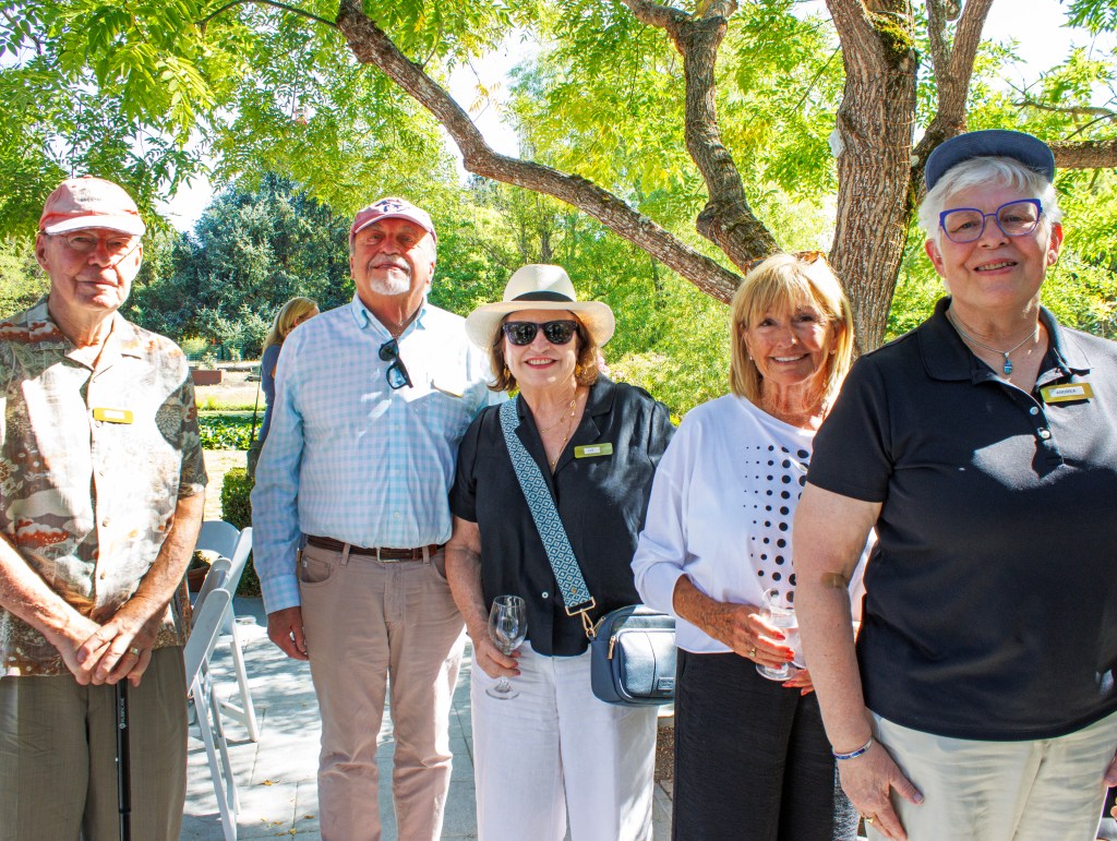 Five older adults standing outside in the shade, surrounded by a tree, a scenic background, and scattered chairs, all smil...