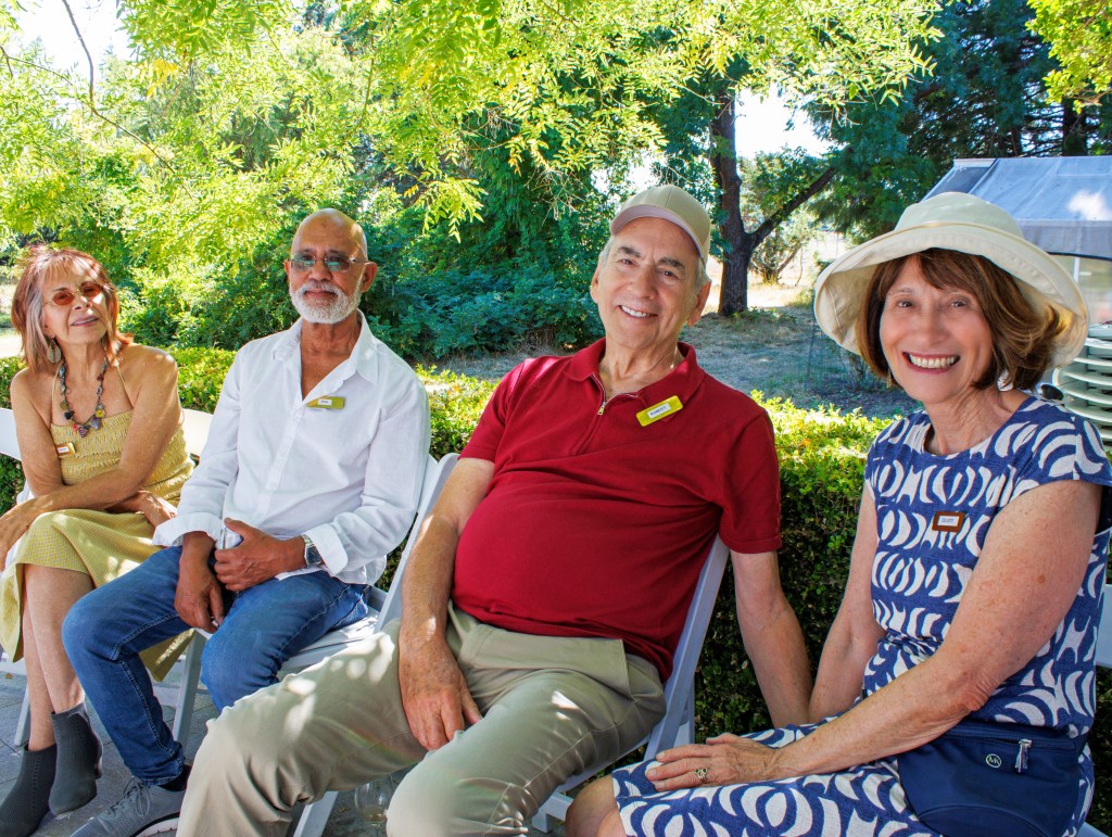 The image shows four middle-aged people sitting in a row, on blue chairs. They are positioned centrally, with a green hedg...