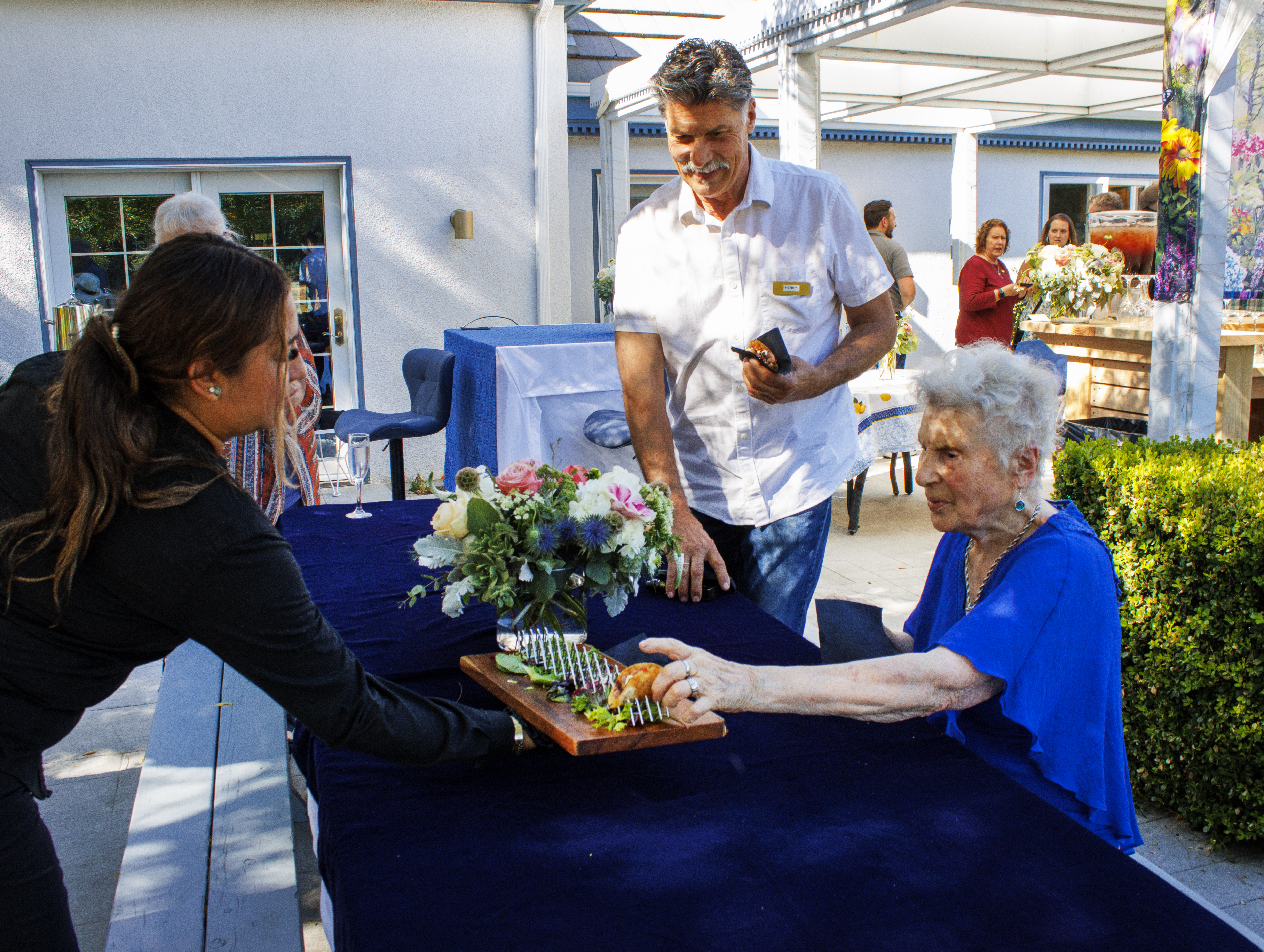 A woman, dressed in a black shirt and sporting long brown hair, reaches for a tray of food from an elderly lady seated at ...