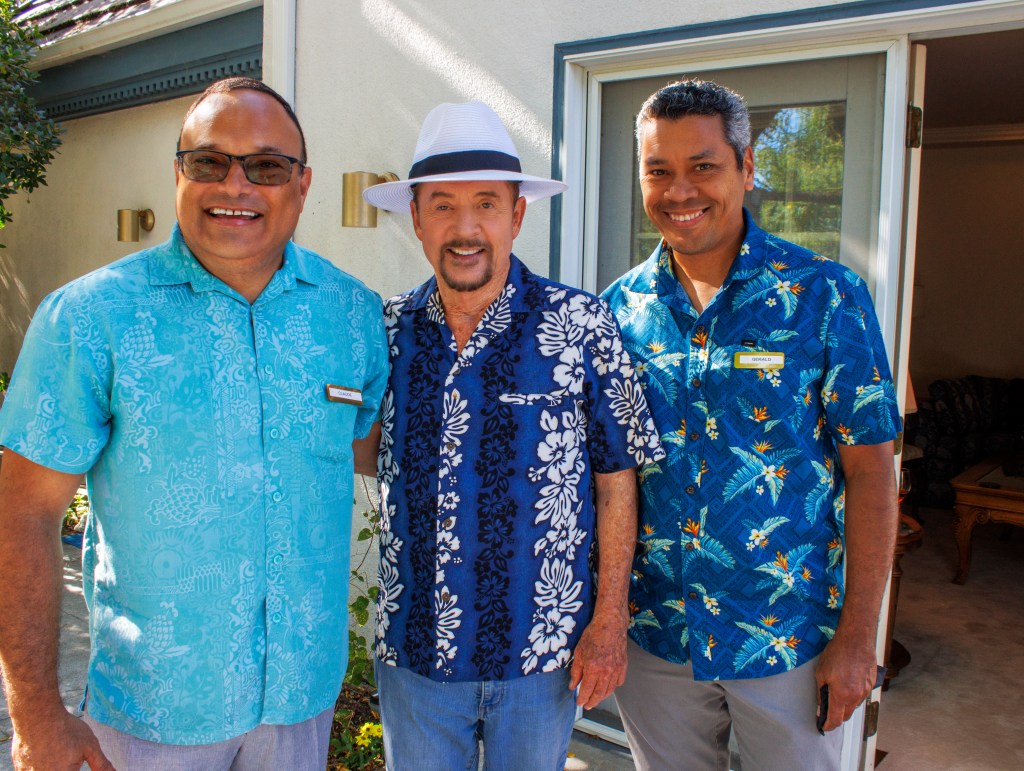 Three men in tropical print shirts standing outside near a sliding door in front of a cream-colored building.