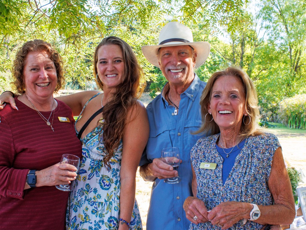 Four people posing for a photo, with one man holding a drink, set against a tree with green leaves and a sunny background.