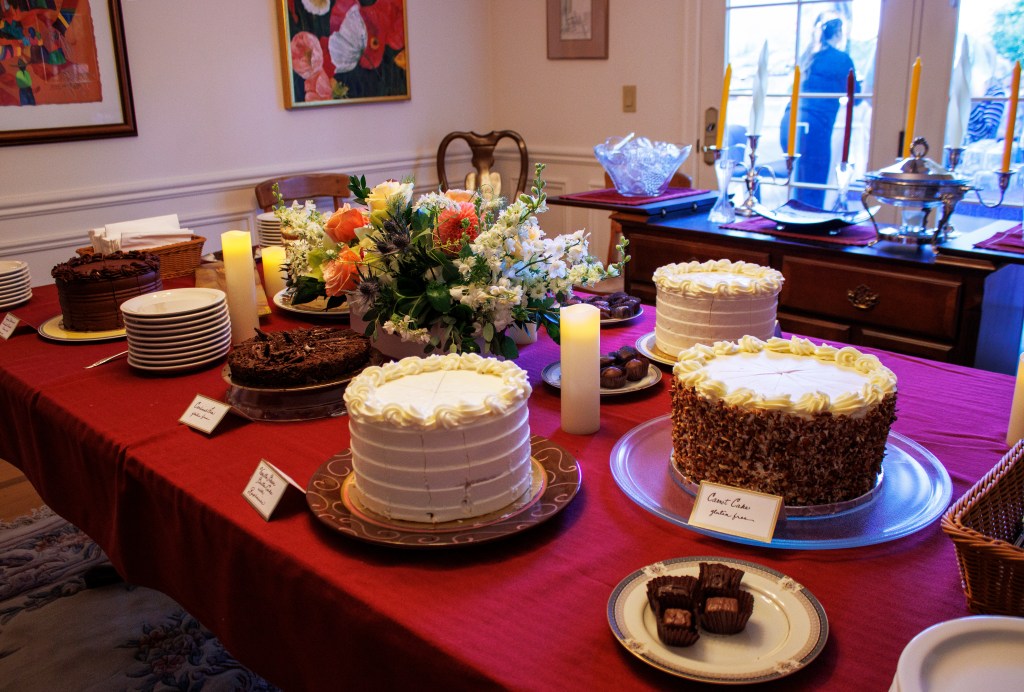 A variety of cakes, chocolate desserts, and flowers are pictured on a table adorned with red tablecloths and white walls f...