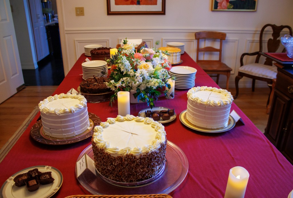 This photograph depicts a long table lavishly adorned with an assortment of plate after plate of decadent cakes and sweet ...
