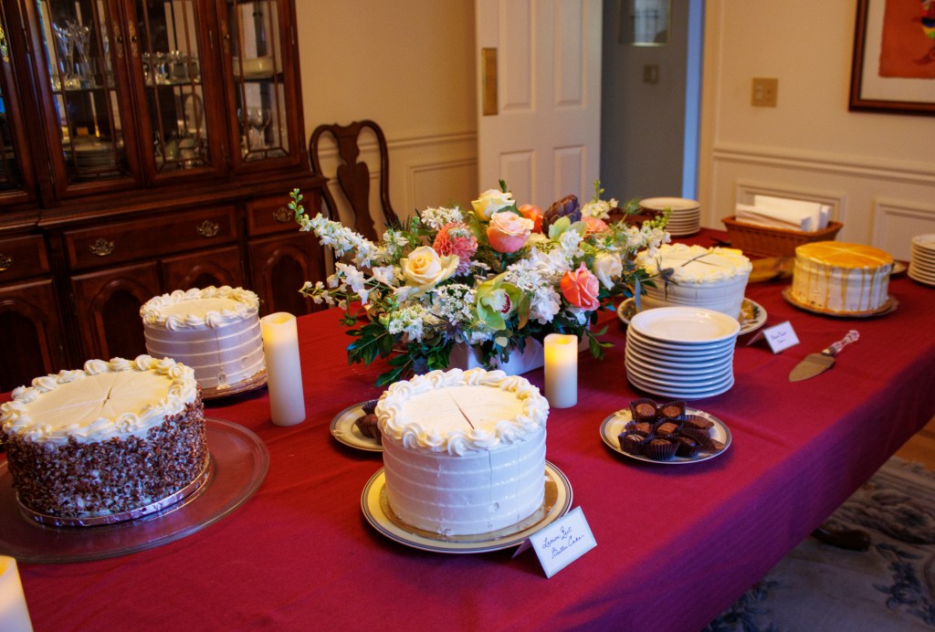 A lengthy table covered in red fabric, featuring a stack of white plates and an assortment of cakes, pies, and flowers.