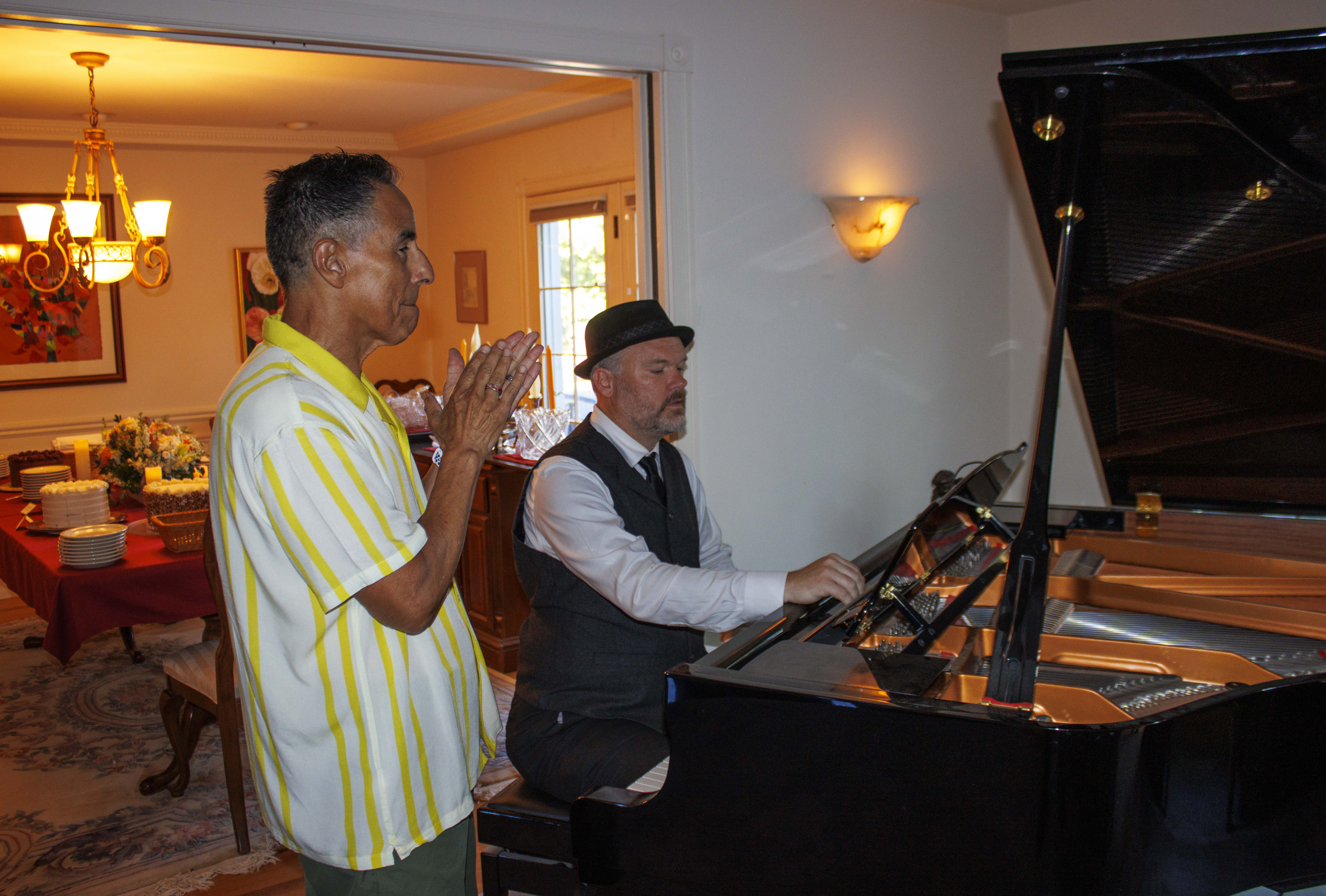 An older Asian man watches while a Caucasian man performs piano. He has short black hair. He is wearing a yellow and white...