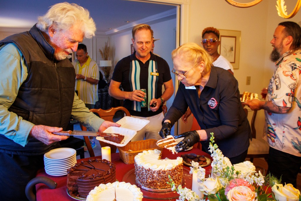 Group of people standing at a dining room table with an assortment of colorful cakes to celebrate a special occasion. A di...