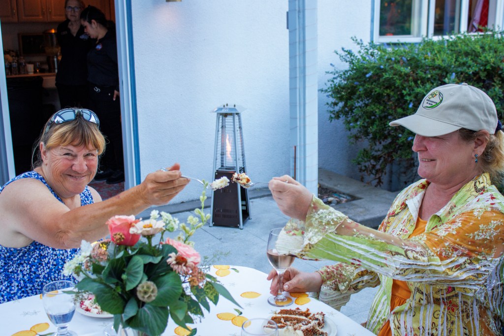 Two women eating cake and outdoor dining. The background is a house and yard.
