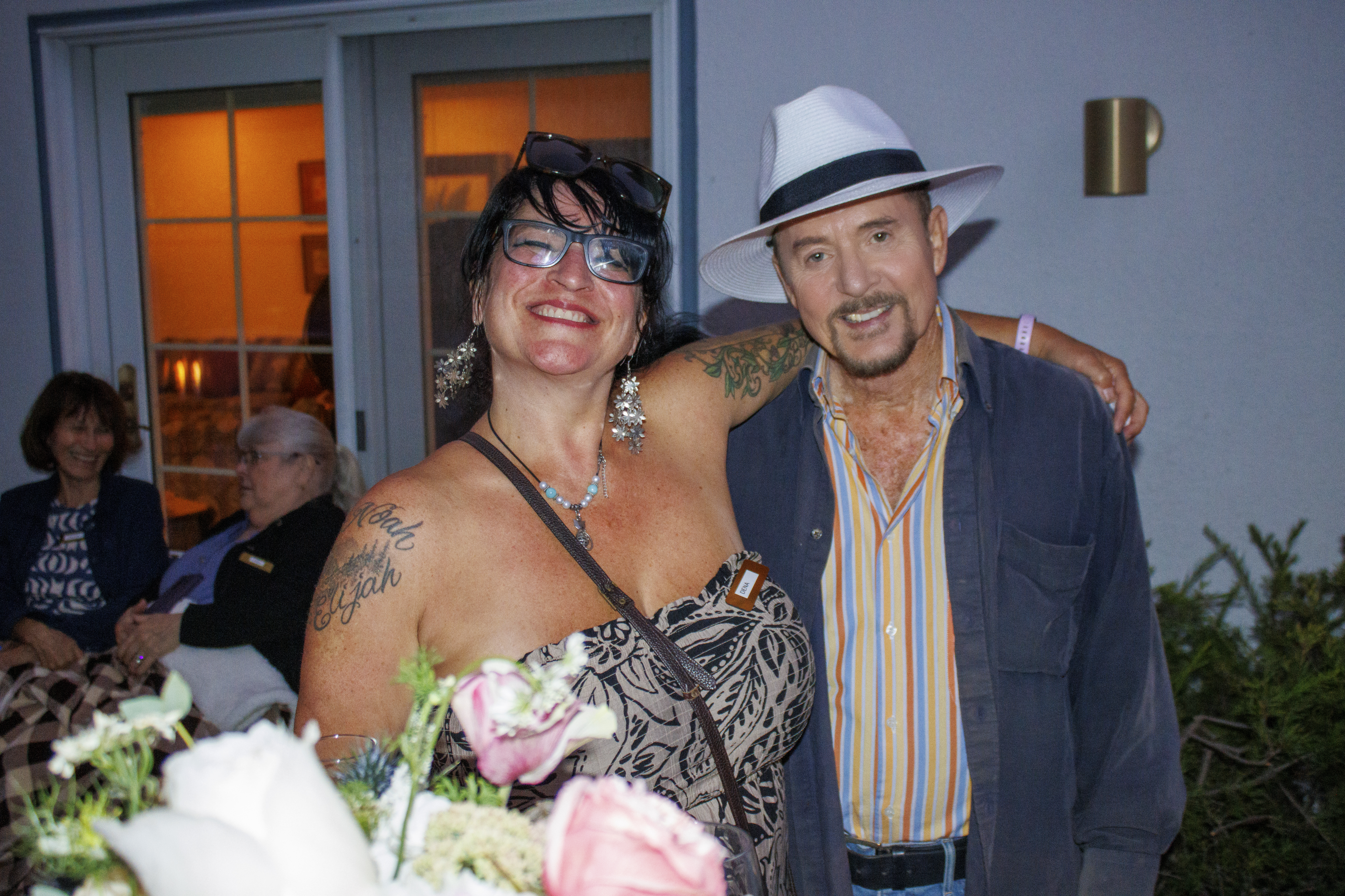 A photograph of a man and woman posing for a picture, standing outside with a group of other people behind them. They appe...